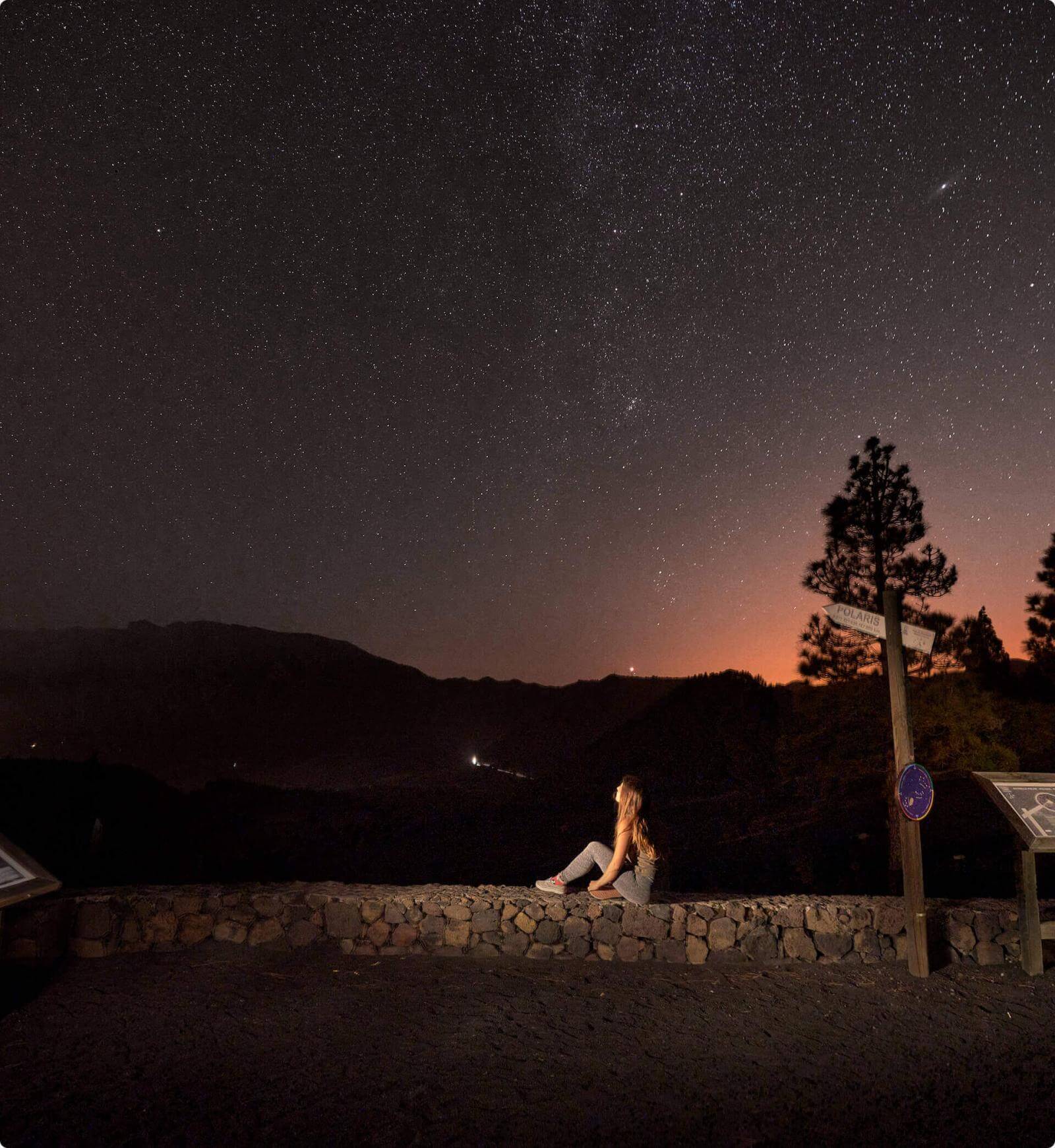 Noite estrelada do miradouro Llano del Jable, em La Palma, com silhuetas de montanhas e uma pessoa.