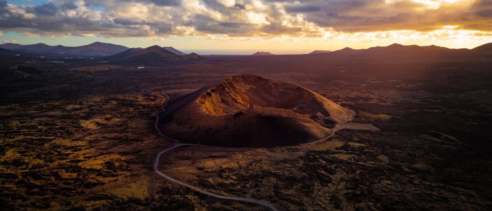 Vista aérea do Vulcão de El Cuervo em Lanzarote, com a cratera iluminada ao pôr-do-sol e paisagem vulcânica.