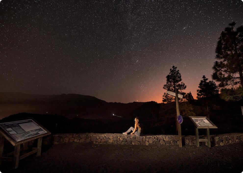 Noite estrelada do miradouro Llano del Jable, em La Palma, com silhuetas de montanhas e uma pessoa.