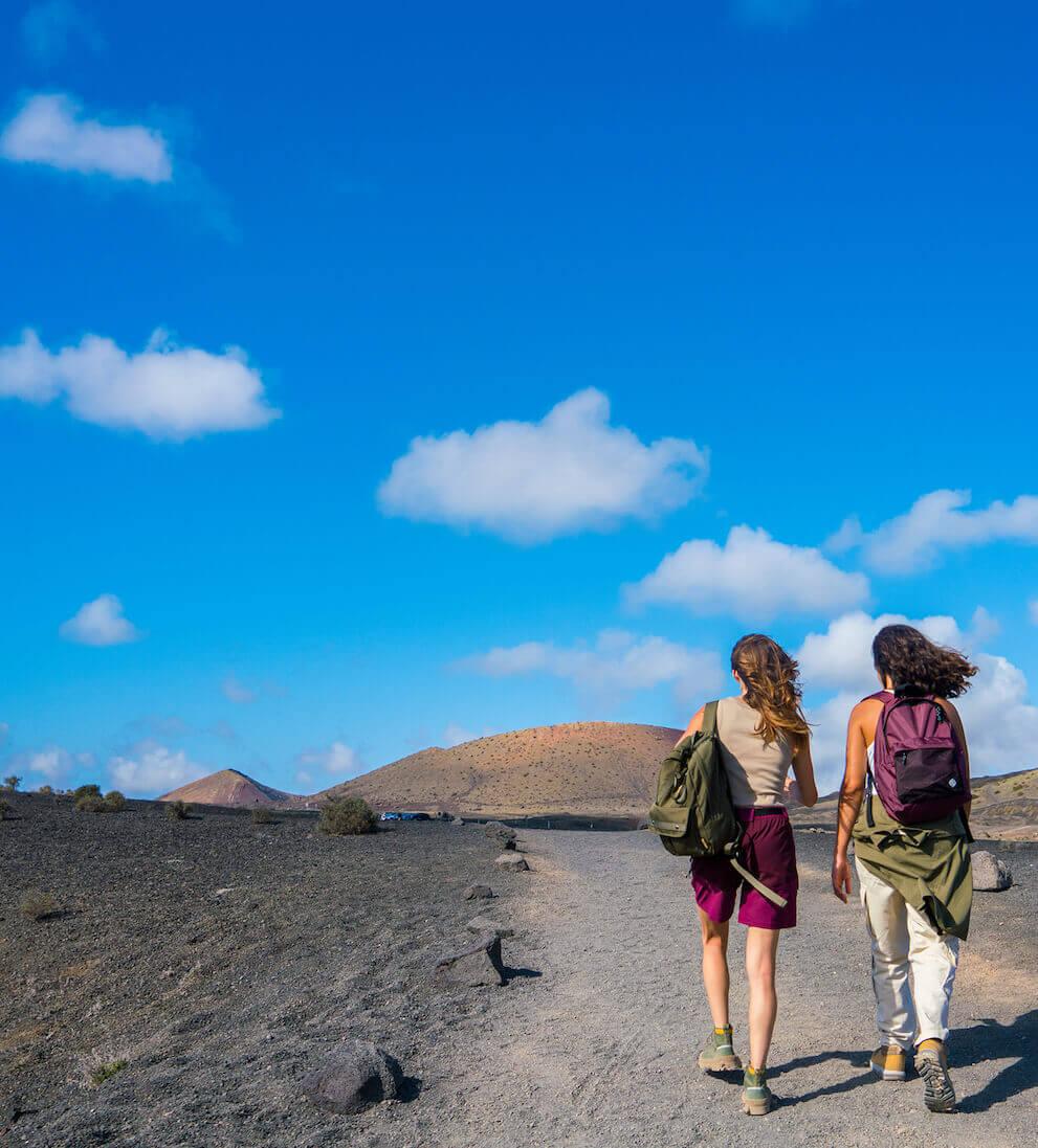 Duas pessoas a andar por um caminho vulcânico no Parque Nacional de Timanfaya sob um céu azul.