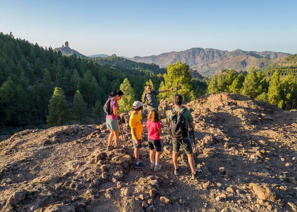 Excursão em grupo pelo Parque Rural do Roque Nublo, entre solo rochoso, pinhal canário e o pico ao fundo.