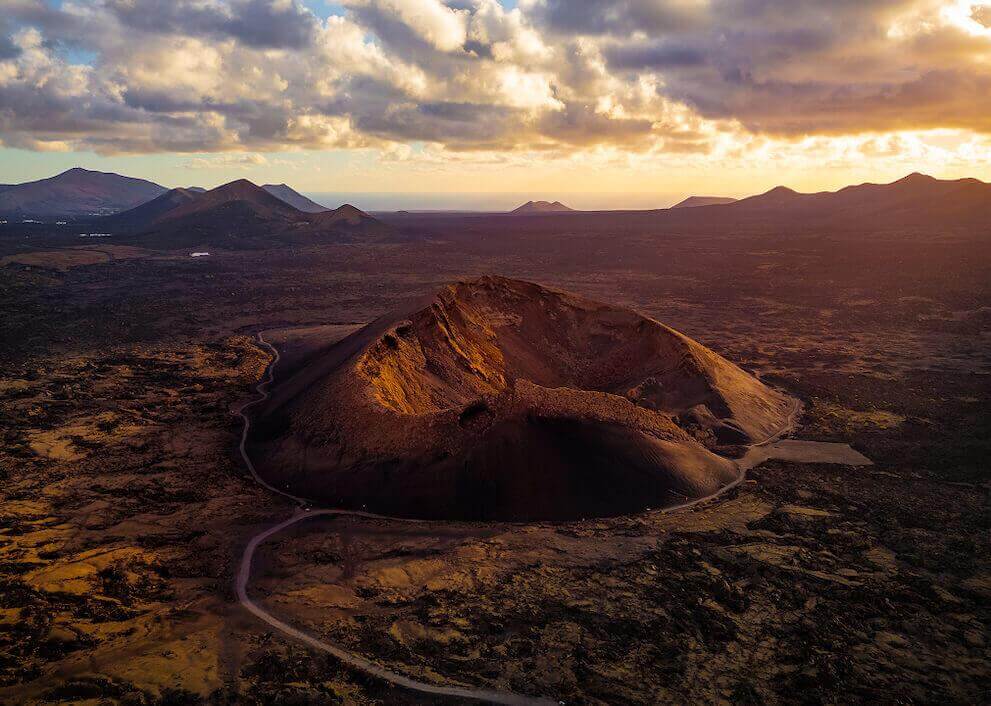 Vista aérea do Vulcão de El Cuervo em Lanzarote, com a cratera iluminada ao pôr-do-sol e paisagem vulcânica.